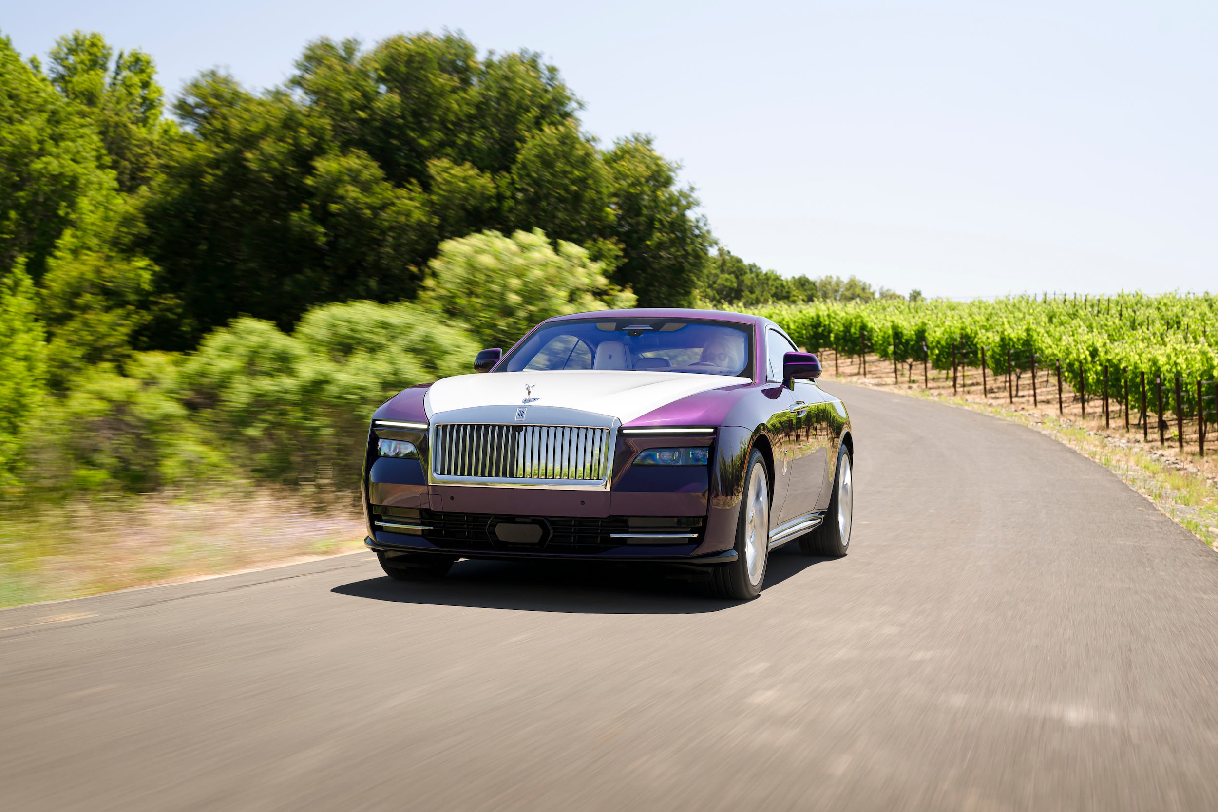 A purple and white Rolls-Royce Spectre car drives down a road with trees in the background.
