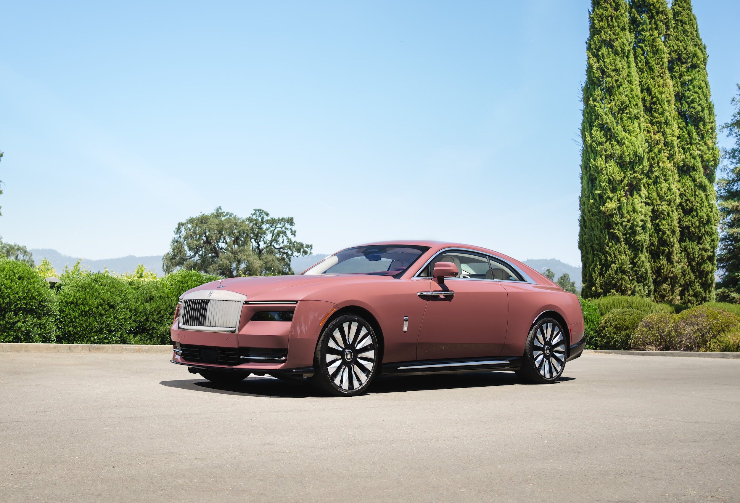 A pink Rolls-Royce Spectre in a parking lot, with blue skies and trees in the background.